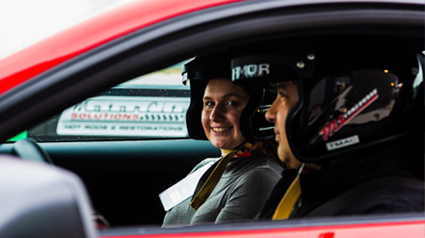 2 people sitting in the front seat of a red car with helmets on, smiling